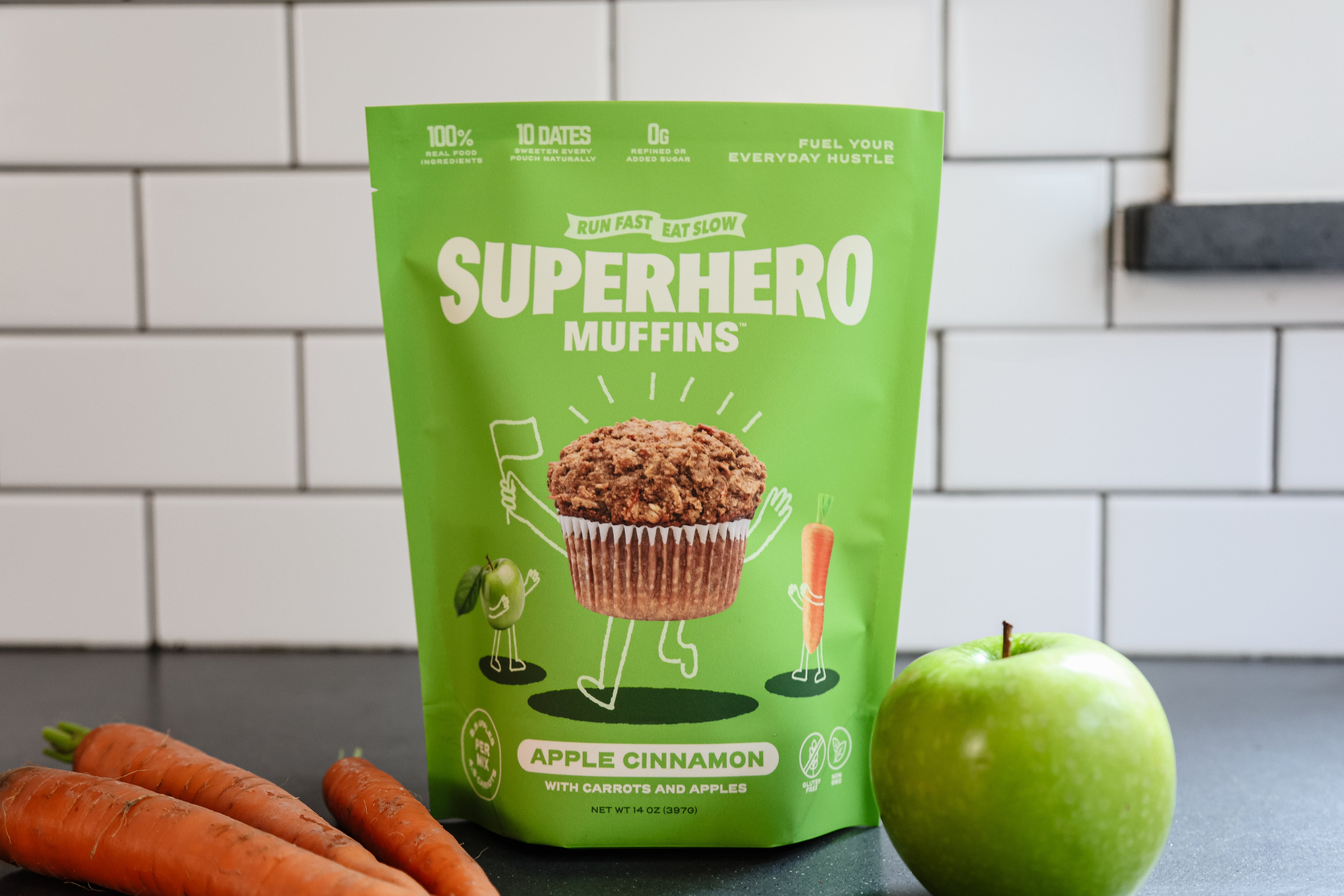 A bag of Superhero Muffin mix on a counter with carrots and an apple.