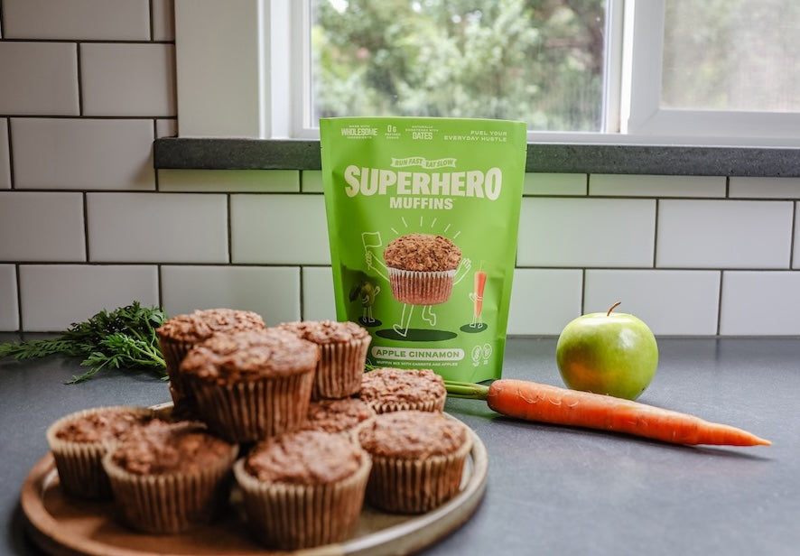 A bag of Superhero Muffin mix on a counter with carrots and an apple next to a plate of Superhero Muffins.