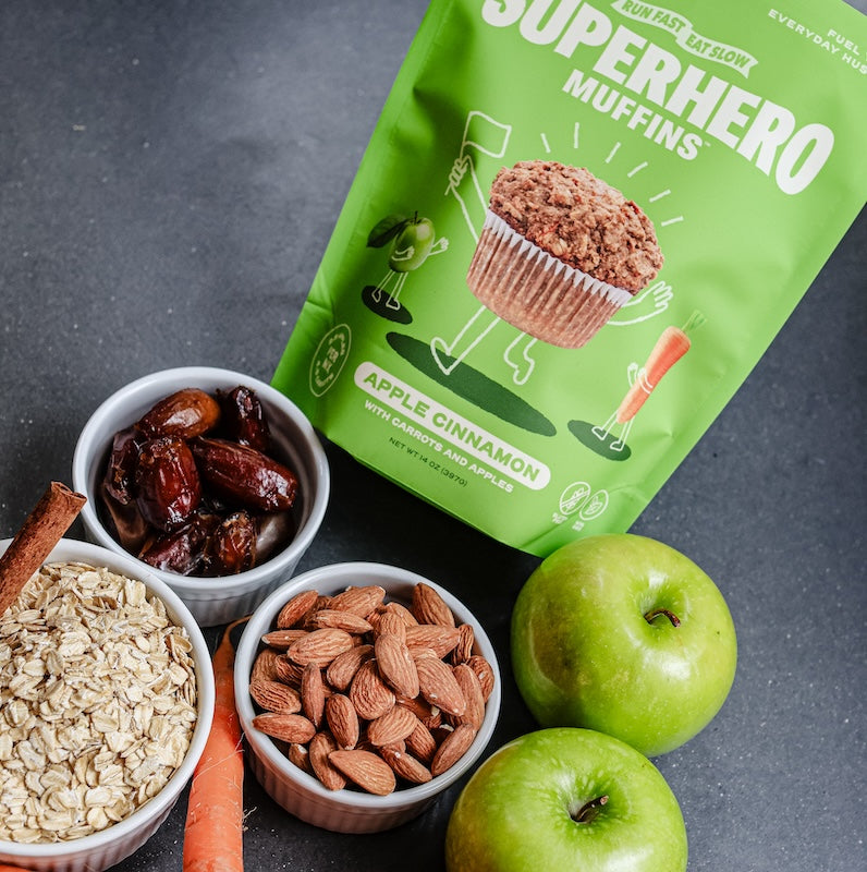 A green bag of Superhero Muffins Apple Cinnamon Muffin Mix on a counter with bowls of dates, almonds, and oats.