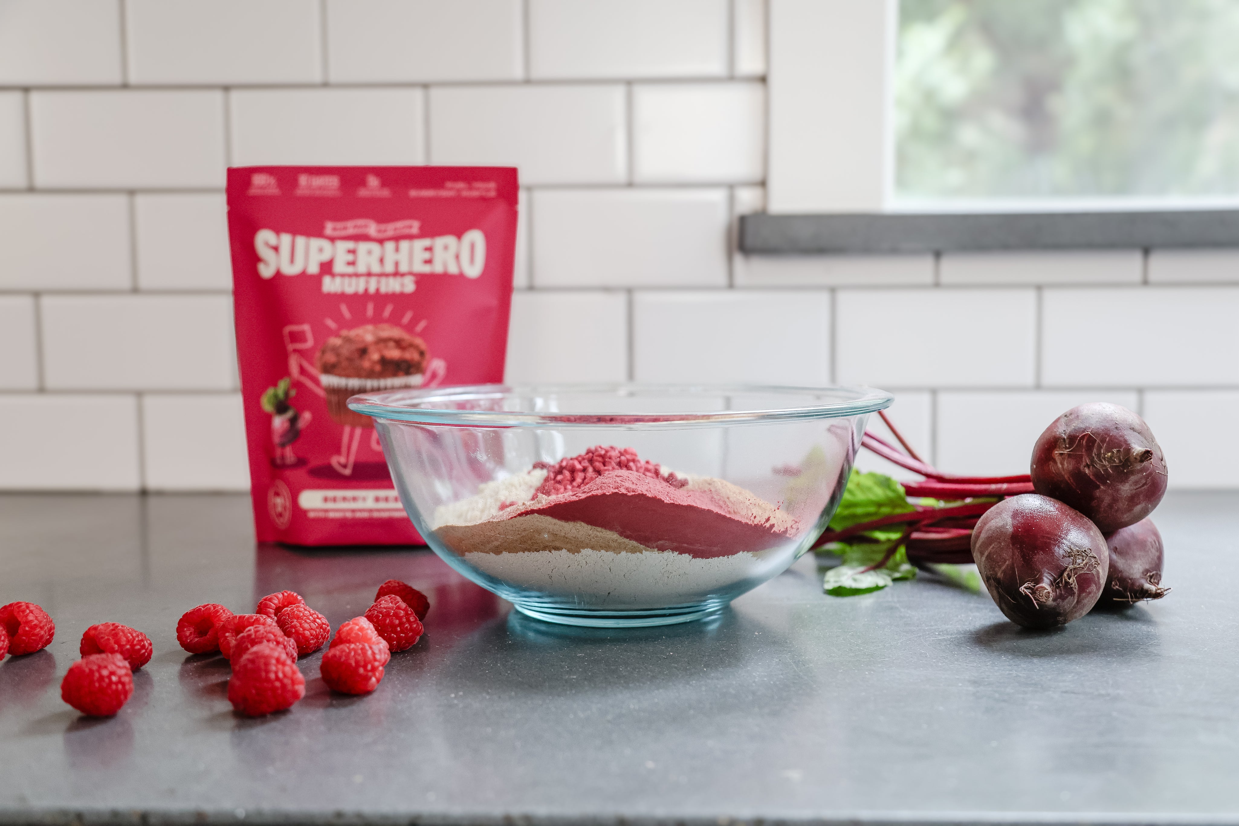 Superhero Muffins Berry Beet Muffin Mix packaging on a kitchen counter with berries, beets, and a bowl of mix.