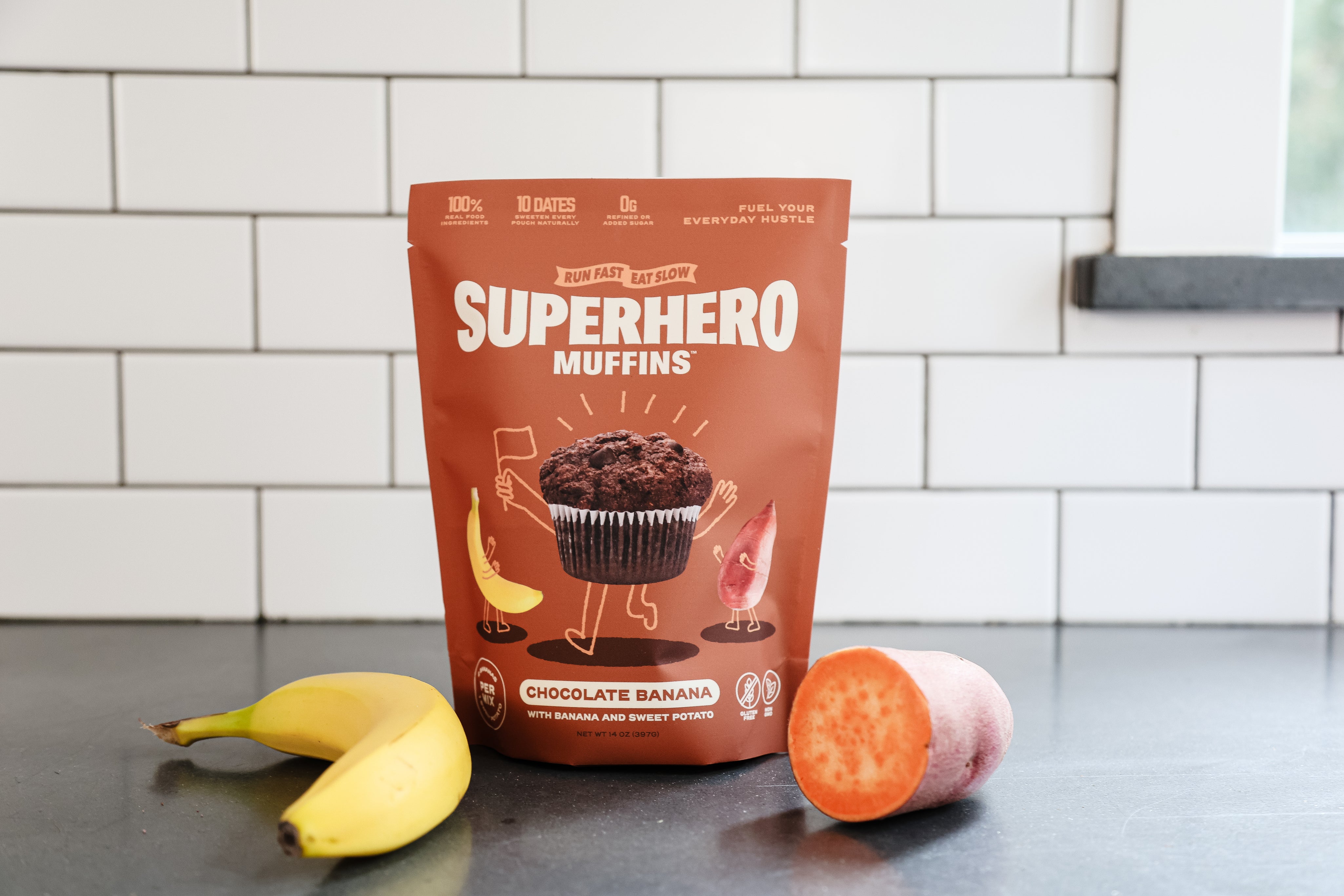 A bag of Run Fast. East Slow. Superhero Muffin mix on a counter with a banana and sweet potato