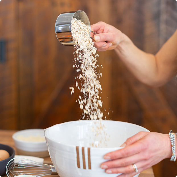 Superhero Muffins Founder Elyse Kopecky pouring oats from a measuring cup into a white bowl on a wooden surface.