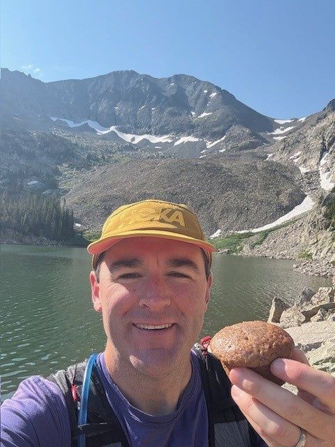 Superhero Muffins customer JP Robb wearing a yellow hat and a backpack holding a muffin with a lake and mountains in the background.